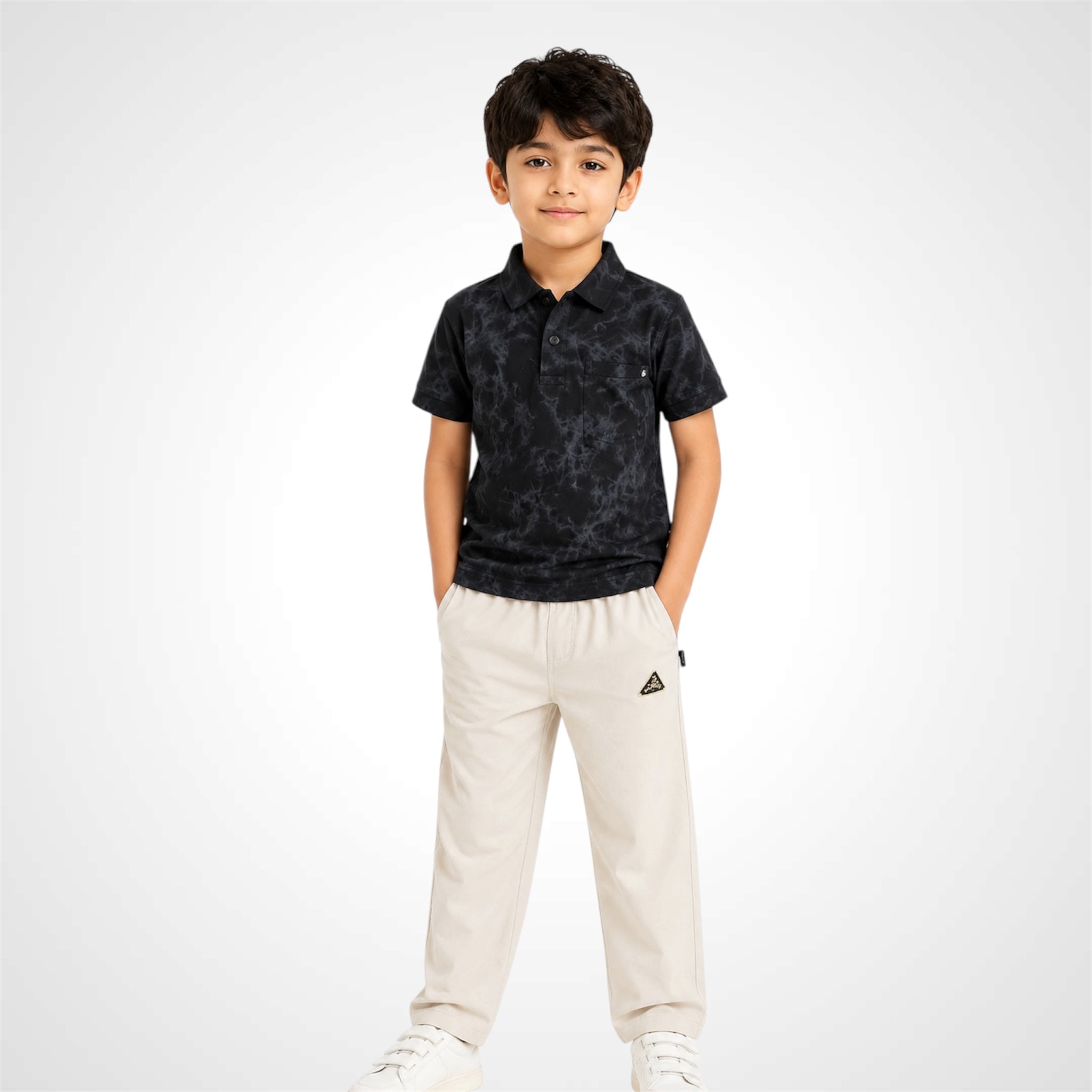 A young boy modeling a short-sleeve polo shirt with a dark gray and black abstract pattern, paired with off-white trousers and white sneakers, set against a light gray studio background.