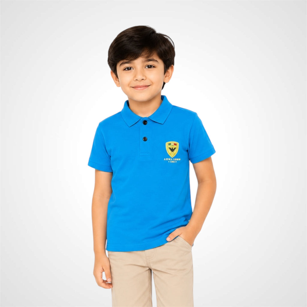 A young boy wearing a bright blue polo shirt with a yellow and black shield logo on the left chest, paired with beige trousers, standing against a light gray studio background.