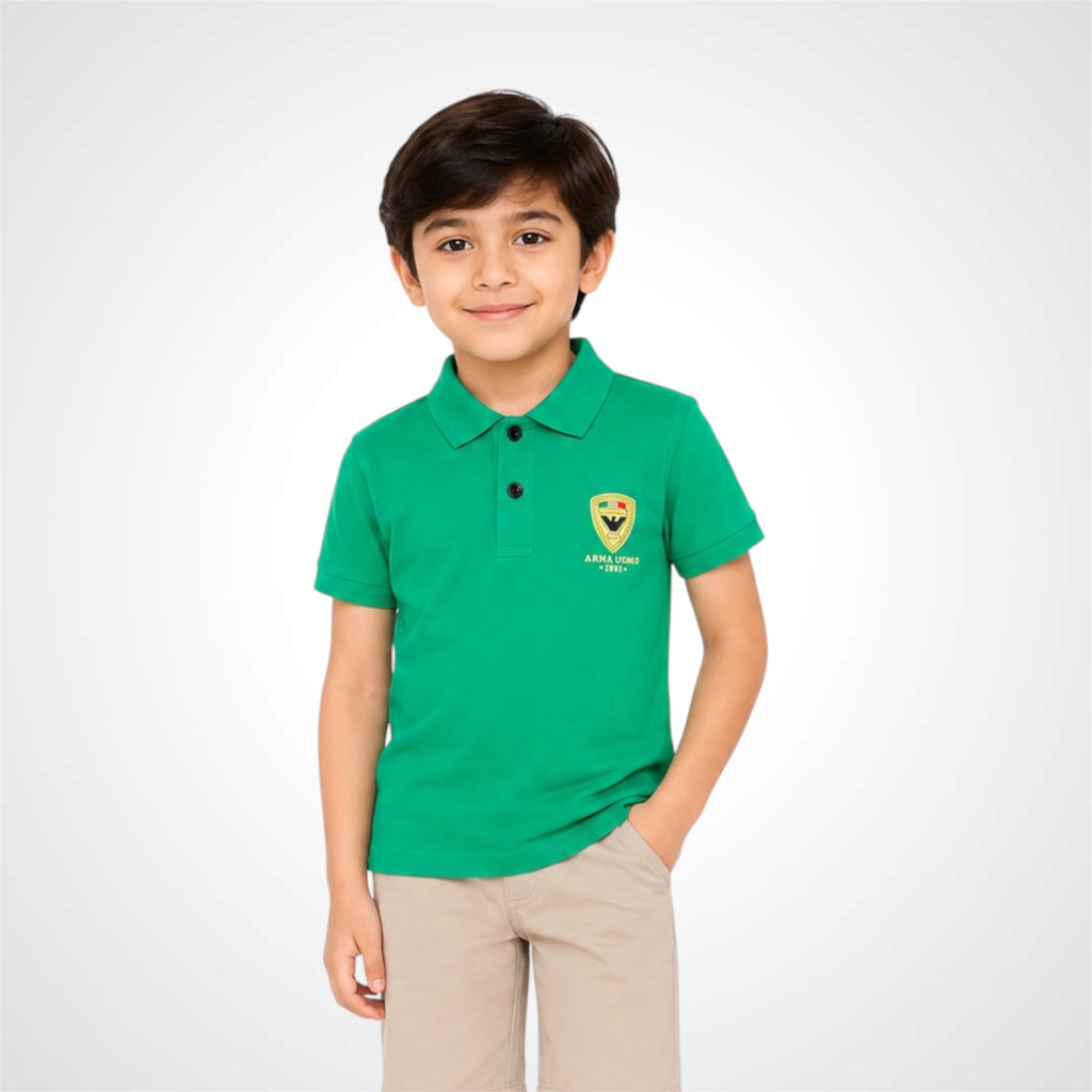 A young boy wearing a bright green polo shirt with a yellow crest logo on the left chest, paired with beige trousers, standing against a light gray studio background.