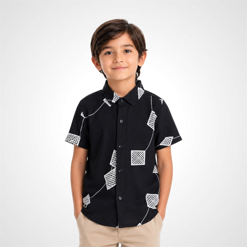 A young boy wearing a black short-sleeve button-up shirt with a white geometric pattern, paired with khaki pants, standing against a light gray studio background.