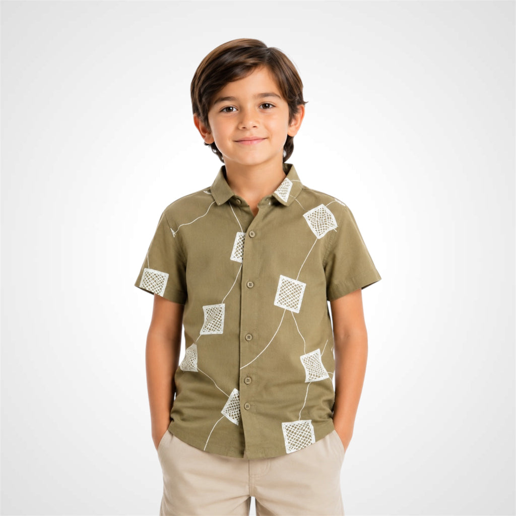 A young boy wearing a short-sleeve button-up shirt in olive green with white geometric patterns, paired with beige pants, standing against a light gray studio background.