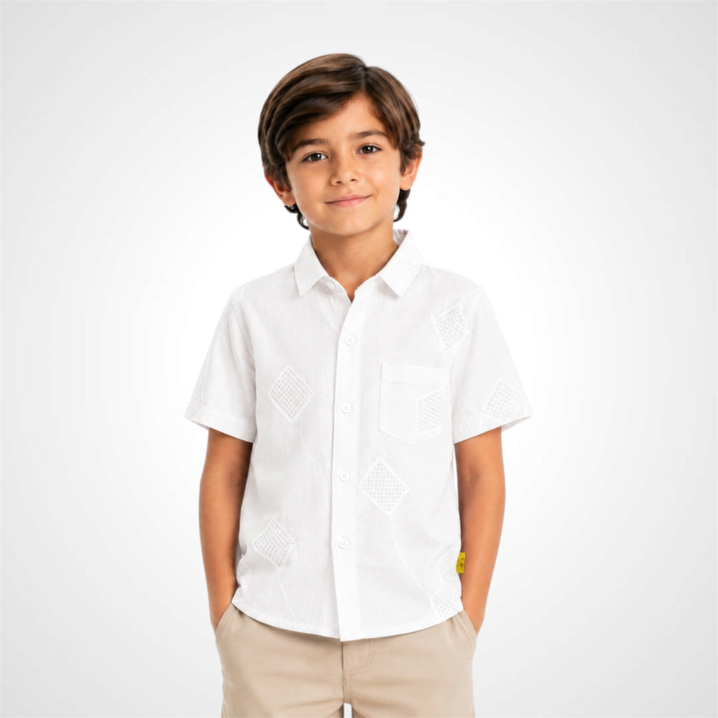 A young boy wearing a white short-sleeve button-up shirt with geometric embroidery on the front and beige pants, standing against a light gray studio background.