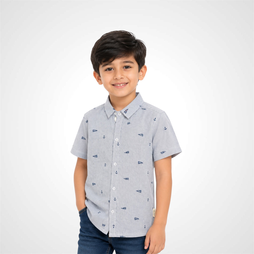 A young boy smiling and wearing a light gray short-sleeve button-up shirt with a small navy blue nautical pattern, paired with dark blue jeans, against a light gray studio background.