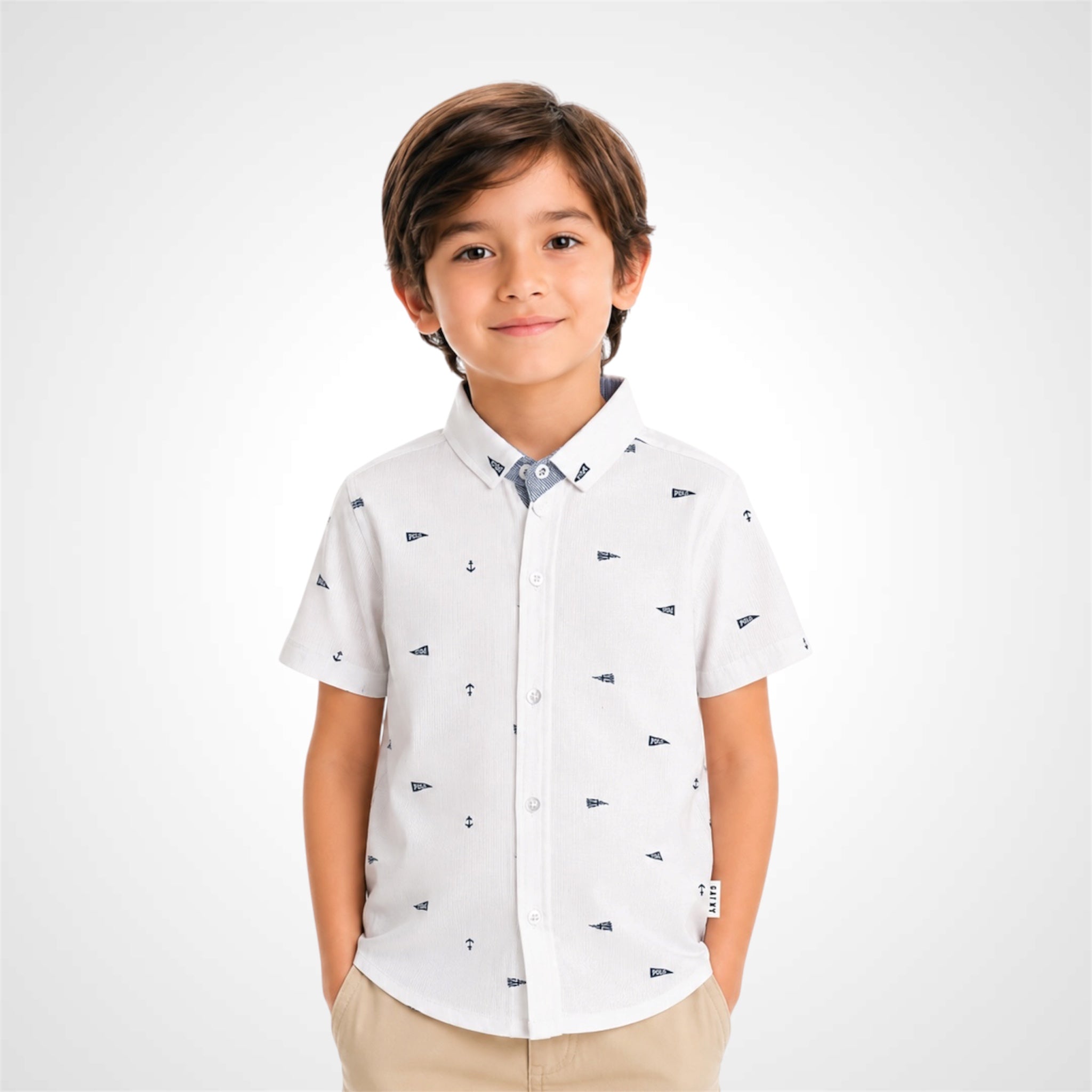 A young boy wearing a white short-sleeve button-up shirt with a small navy blue anchor pattern, paired with beige trousers, standing against a light gray studio background.