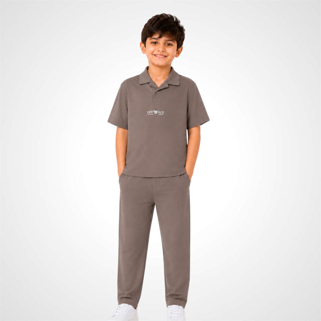 A young boy modeling a matching two-piece school uniform set in a solid taupe-brown color, featuring a short-sleeve polo shirt and matching trousers, against a light gray studio background.